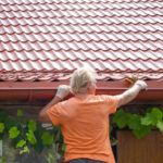 Man in orange shirt stands reaching in the gutters of a red roofed home.