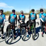 The team of five men stand together, holding their bikes, wearing blue matching riding vests.