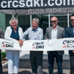 In order, Kyle, Robyn, Cris, Ryan and Matt Douglas stand together smiling and holding a DKI-CRCS ribbon as Cris prepares to cut it