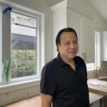 A man in a black shirt stands in a white kitchen, looking at the camera.