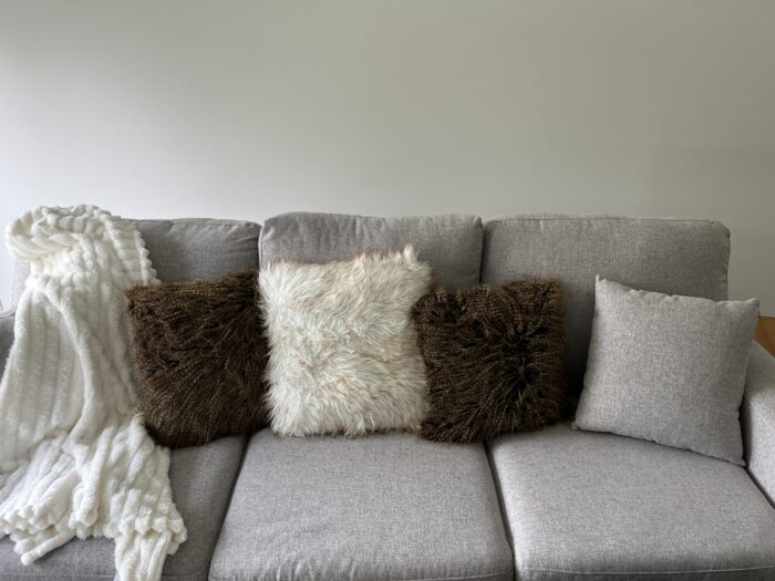 A light grey couch is shown in front of a white wall with two brown fluffy pillows, a grey pillow, a white fluffy pillow and a white throw blanket on the left cushion. 