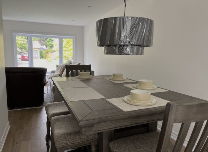 A dining room table with four places set and a low hanging chandelier with the plastic wrap still on it. In the background you see light coming in through the window and the side of the couch.