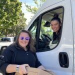 One woman stands beside a white van holding a cardboard box, while another woman sitting in the driver's seat leans out the window with a smile.